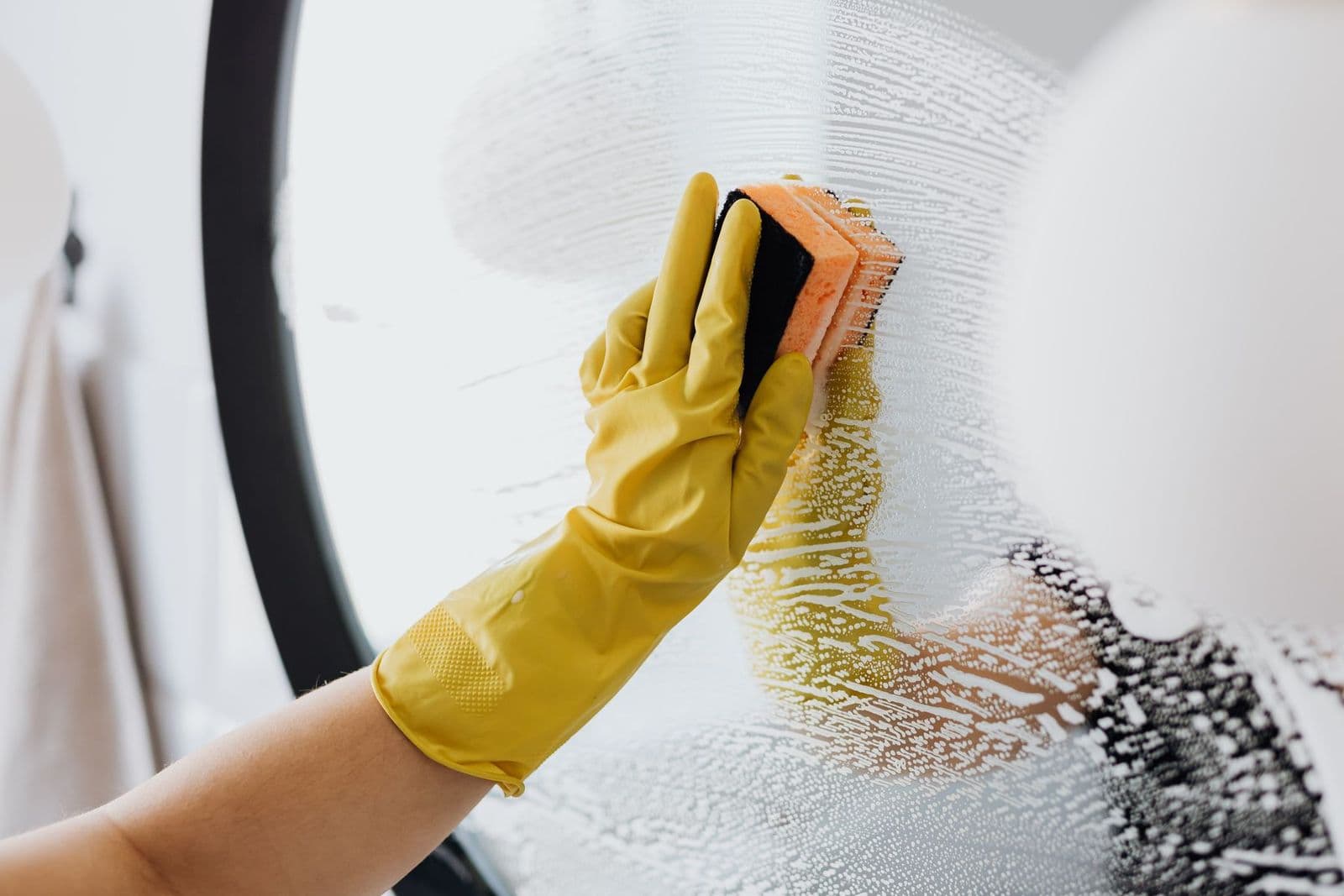 Gloved hand cleaning a mirror with a sponge and soapy water